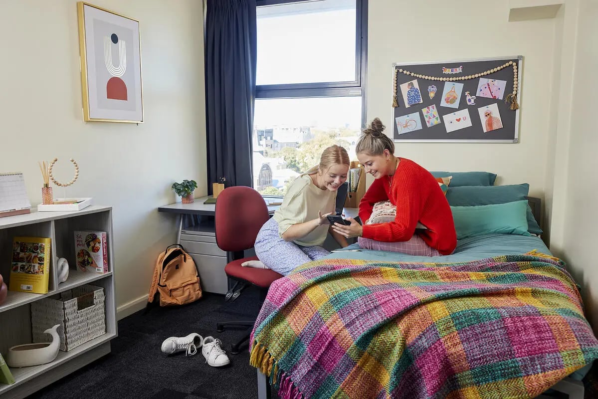 Two students on a colorful bed using phones bedroom at Broadway x Scape in Sydney Student Accommodation