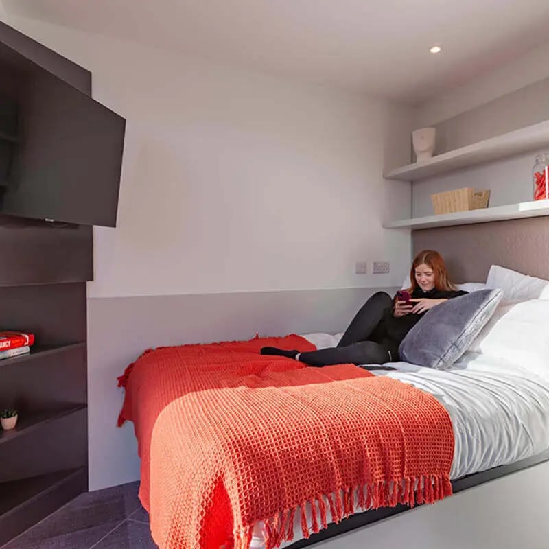 Bed with orange throw student uses phone bedroom at True Glasgow in Glasgow Student Accommodation