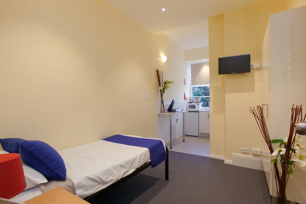 Single bed with blue pillows kitchen area studio apartment at ume 142 Darlinghurst Village in Sydney Student Accommodation