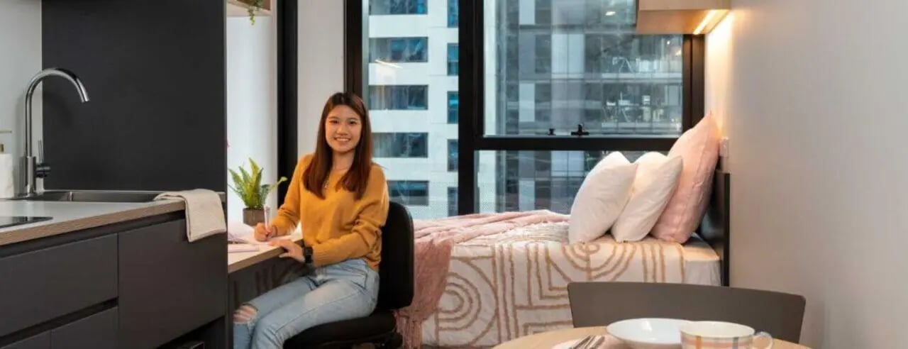 Student studying at desk by bed and kitchen area bedroom at UniLodge Melbourne Central in Melbourne Student Accommodation