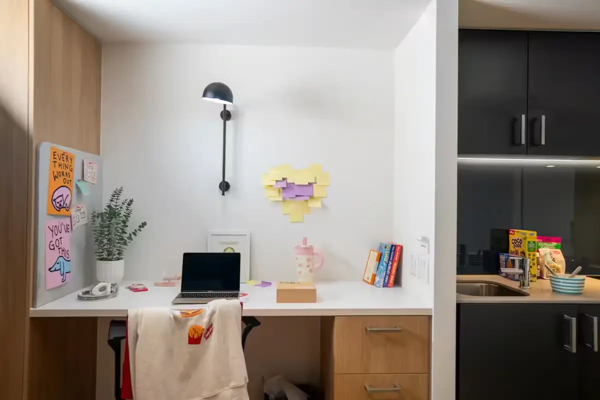 Desk with laptop, plant, books, and kitchen area. at Scape Uni Square in Melbourne Student Accommodation