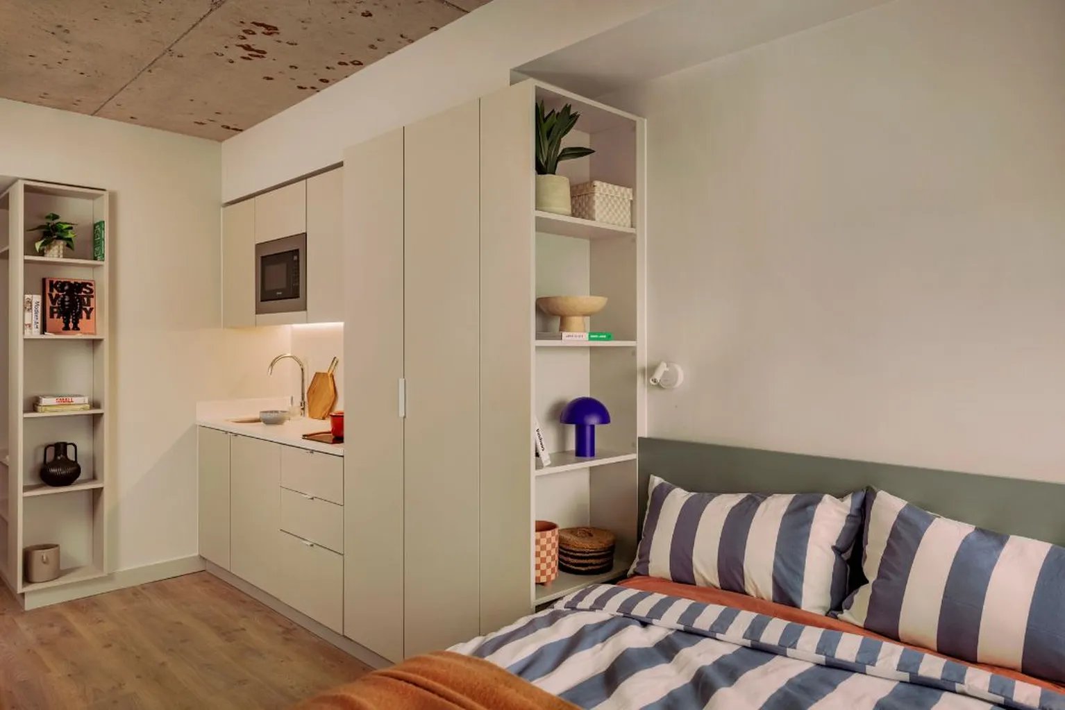 Bed with striped pillows, shelving, and kitchenette bedroom at Scape Hammersmith in London Student Accommodation