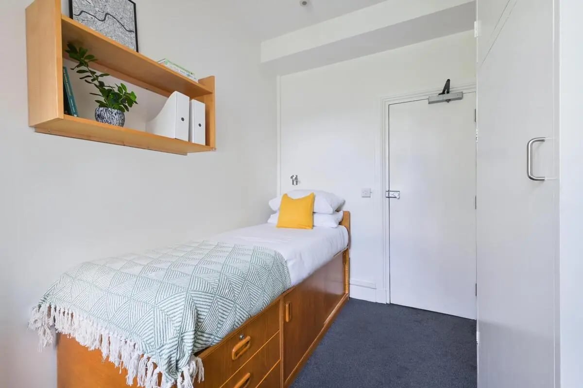 Bed with white bedding yellow pillow shelving bedroom at AXO Waterloo in London Student Accommodation