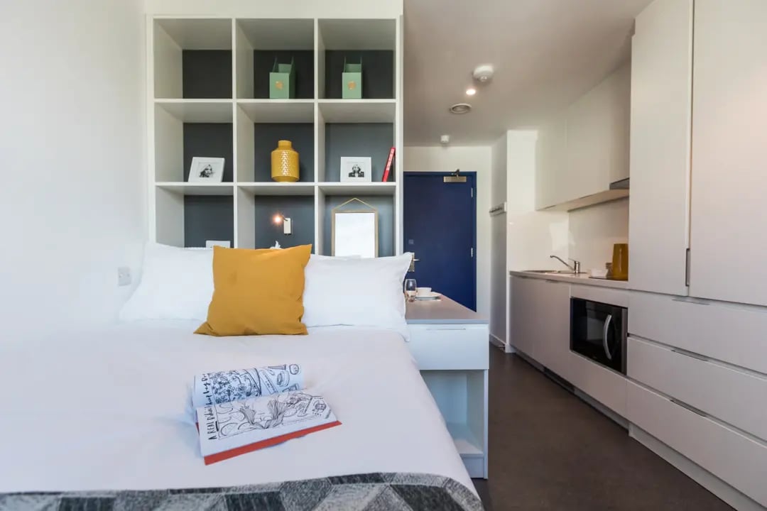 Bed with yellow pillow shelves kitchen and door studio at Vogue Studios in Brighton Student Accommodation