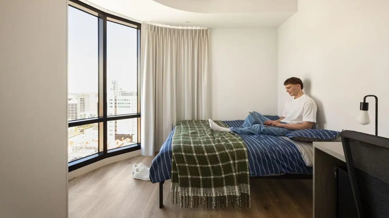 Man on bed with laptop large window bedroom at UniLodge Perth Central in Perth Student Accommodation