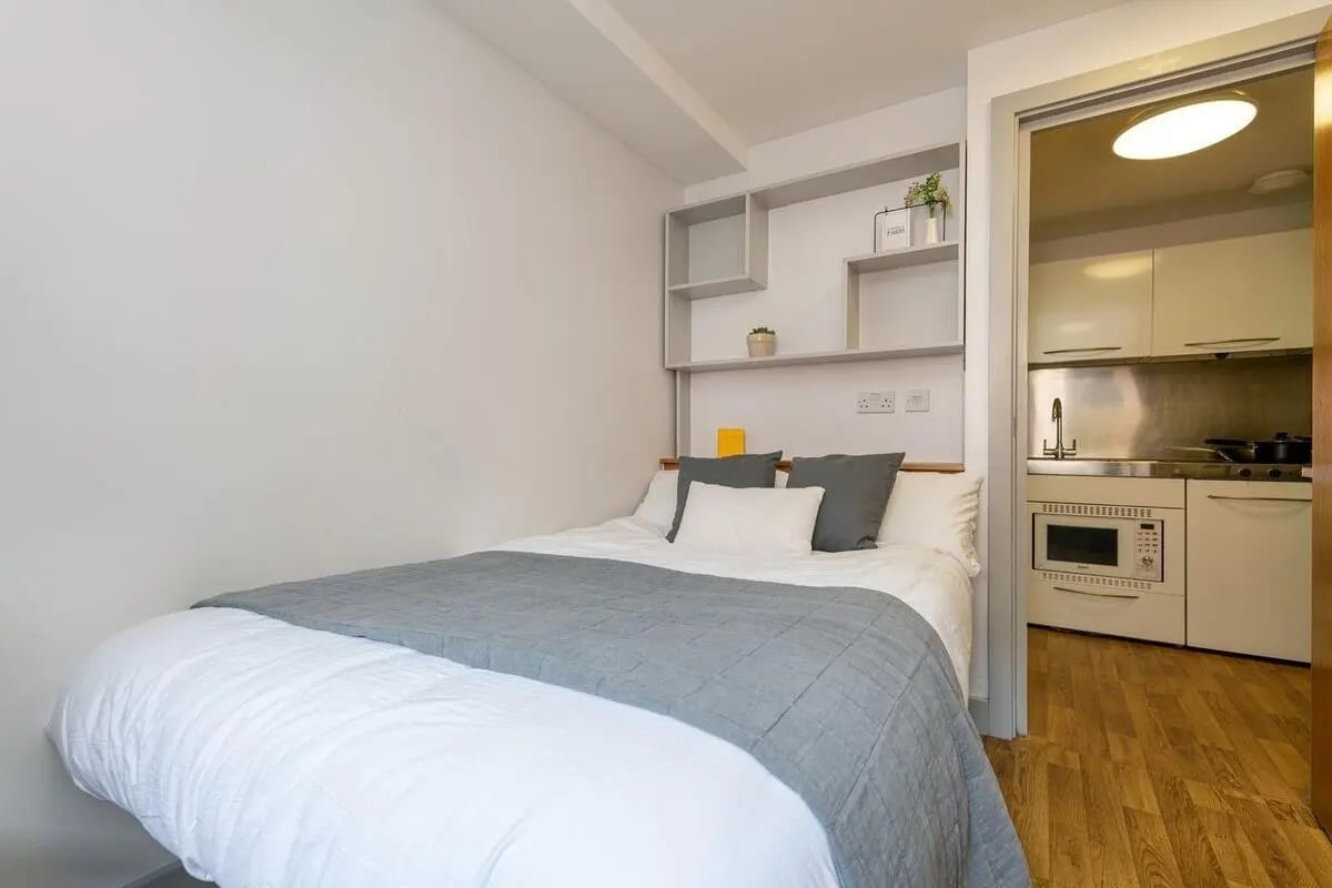 Bed with grey blanket and shelving adjacent kitchen area at Collegiate St James Street in Portsmouth Student Accommodation