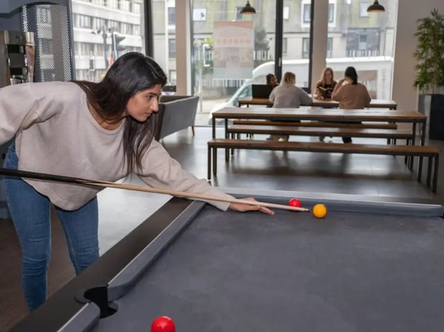 Woman plays pool next to communal study tables and sofas living room at Powis Place in Aberdeen Student Accommodation