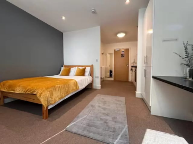 Bed with orange throw grey rug kitchen area bedroom at Hydrogen in Nottingham Student Accommodation