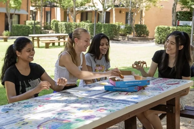 Four smiling students eating pizza outdoor common area at Sydney University Village in Sydney Student Accommodation