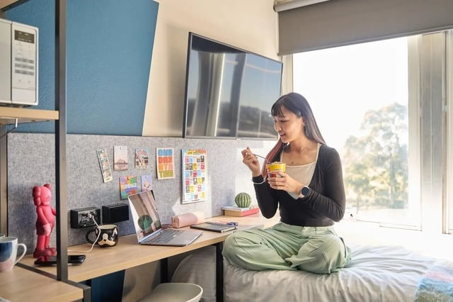 Student eating on bed with desk and TV bedroom at Campus Melbourne in Melbourne Student Accommodation