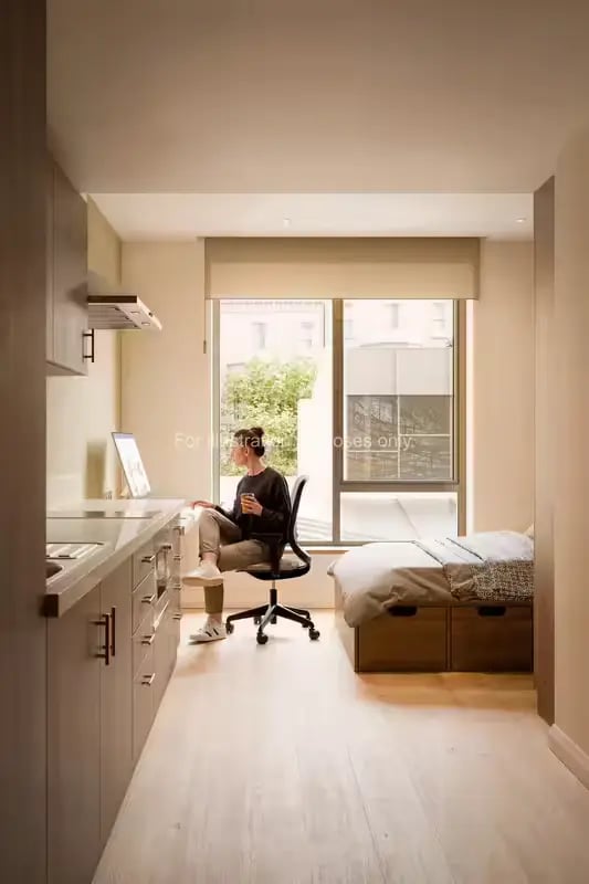 Woman at desk near bed and kitchenette studio apartment at Ryan Hall in Belfast Student Accommodation