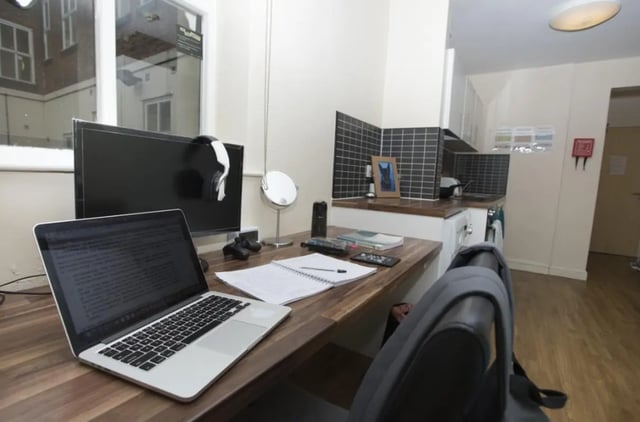 Desk with laptop monitor headphones and kitchen area living room at Daisybank Villas in Manchester Student Accommodation