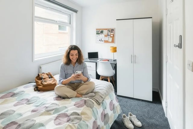 Student using phone on bed with backpack study desk bedroom at Park Village in Sheffield Student Accommodation