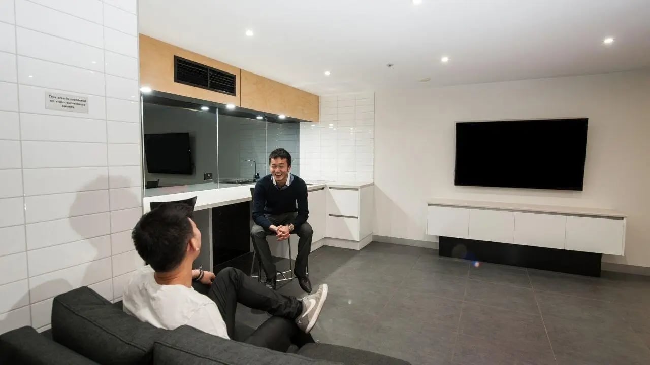 Two men one on sofa kitchen counter TV living room at Student Living on Lonsdale in Melbourne Student Accommodation
