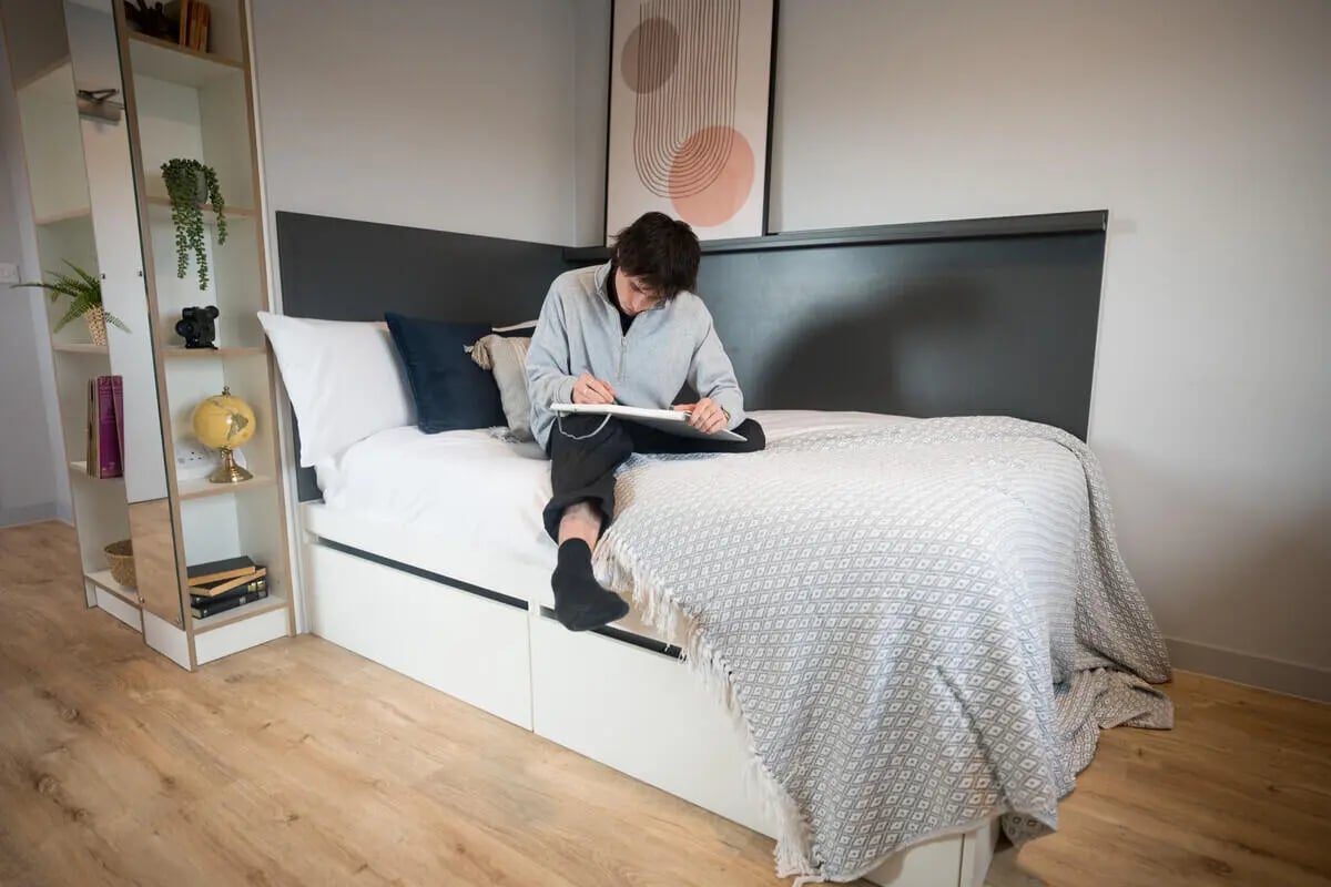 Student writing on bed near bookshelf bedroom at West Wing in Cardiff Student Accommodation