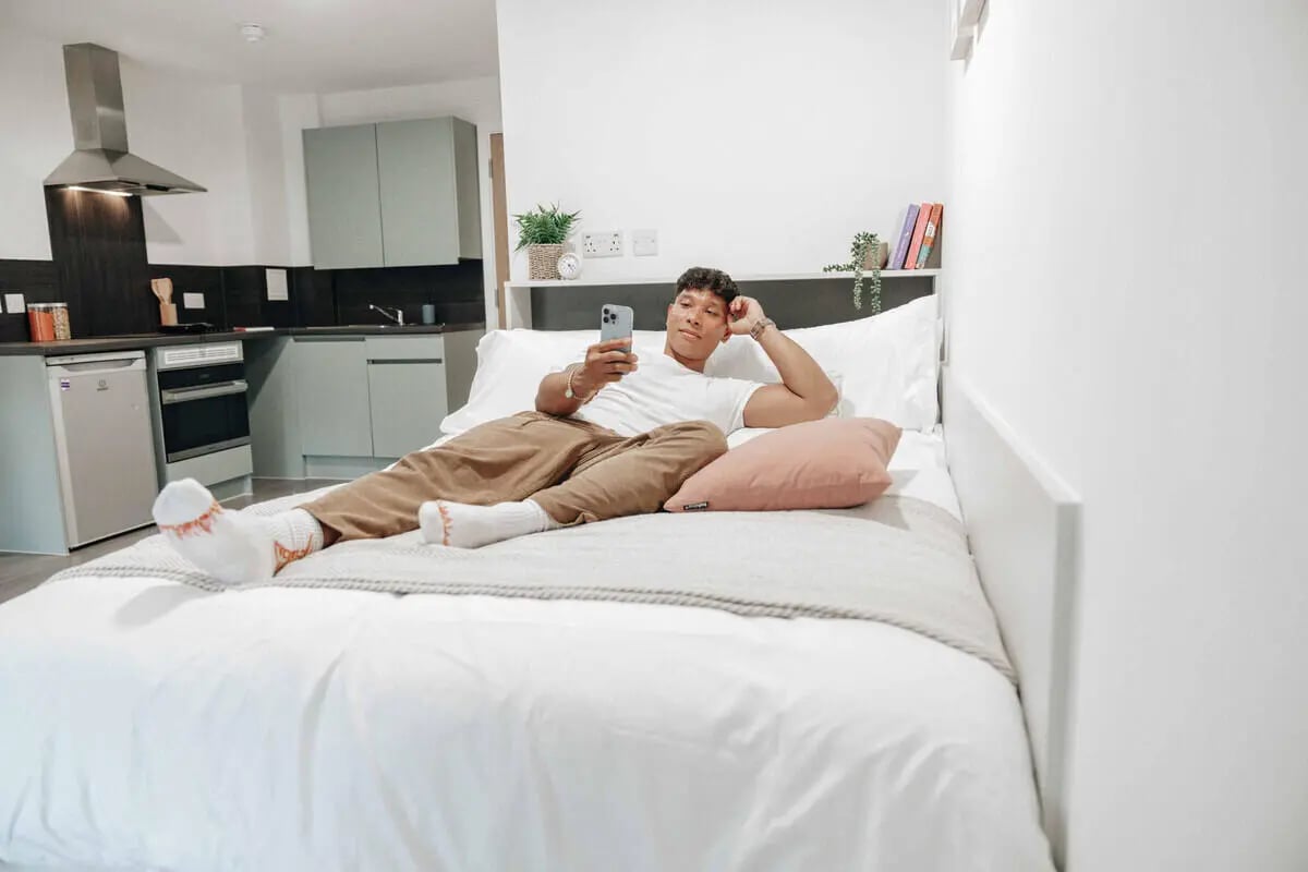 Man on bed with phone kitchen area bedroom at Alma Court in Canterbury Student Accommodation
