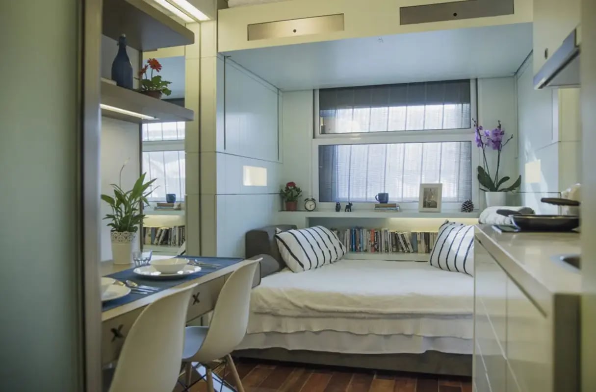 Bed with pillows books desk chairs plant kitchen area at The Stay Club - Camden in London Student Accommodation