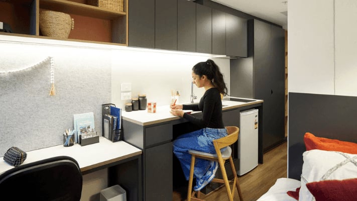 Woman studying at desk with mini fridge and bed. at UniLodge Melbourne Flagstaff in Melbourne Student Accommodation