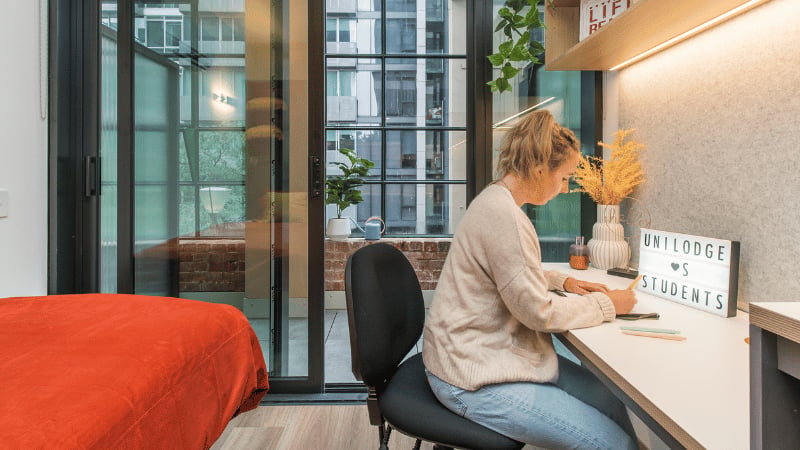 Student studying at desk with orange bed Bedroom study at UniLodge Melbourne Flagstaff in Melbourne Student Accommodation