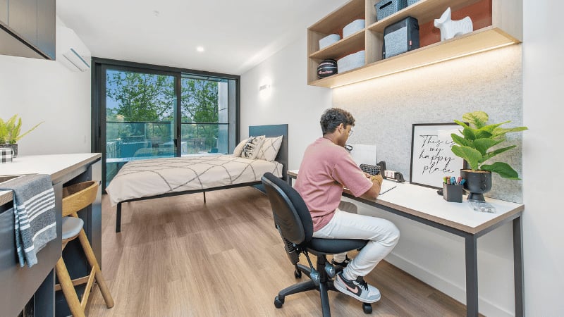 Student at desk with bed kitchen area bedroom at UniLodge Melbourne Flagstaff in Melbourne Student Accommodation