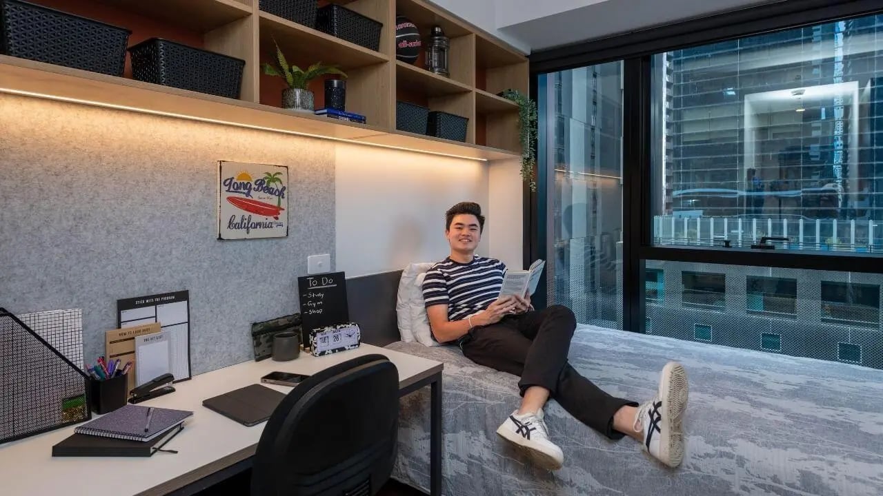 Man relaxing on bed with desk and window bedroom at UniLodge Melbourne Central in Melbourne Student Accommodation