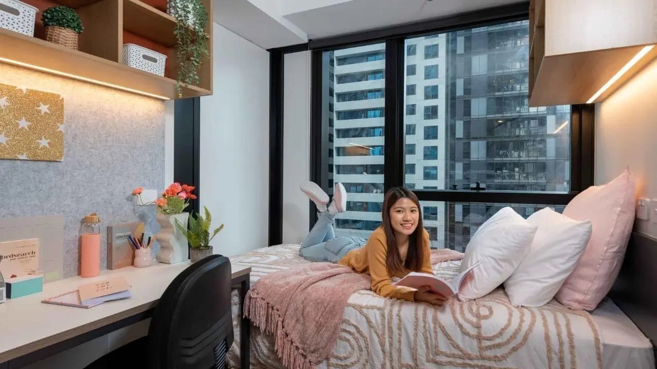 Woman on bed with desk and city view bedroom at UniLodge Melbourne Central in Melbourne Student Accommodation