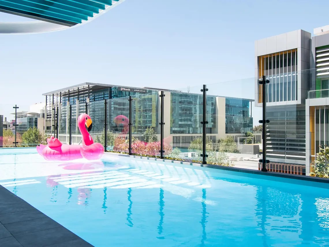 Inflatable flamingos float in a bright blue swimming pool exterior at Yugo Perth City in Perth Student Accommodation
