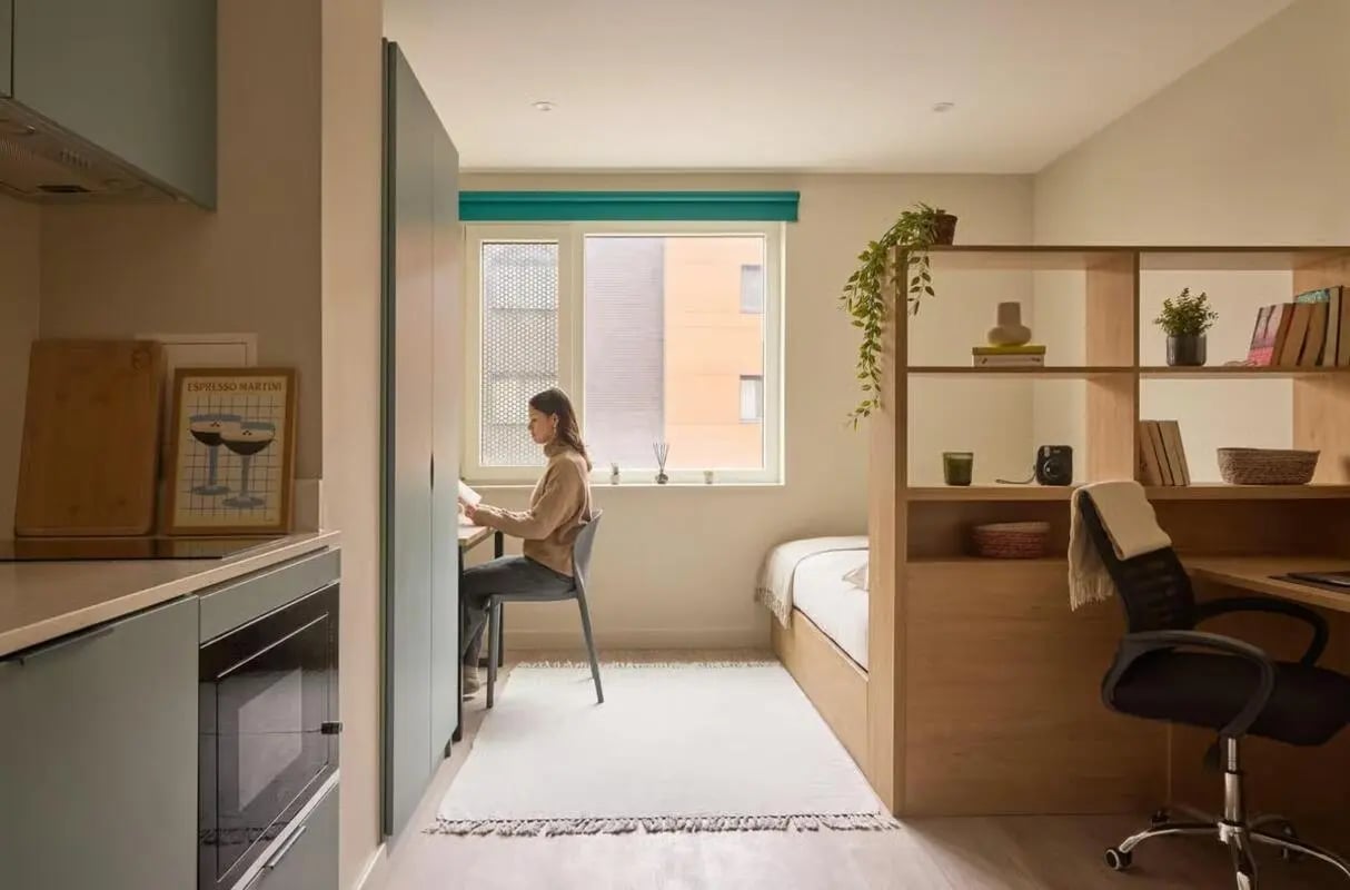 Woman at desk, bed, shelving unit, kitchen Bedroom at Ark Canning Town in London Student Accommodation