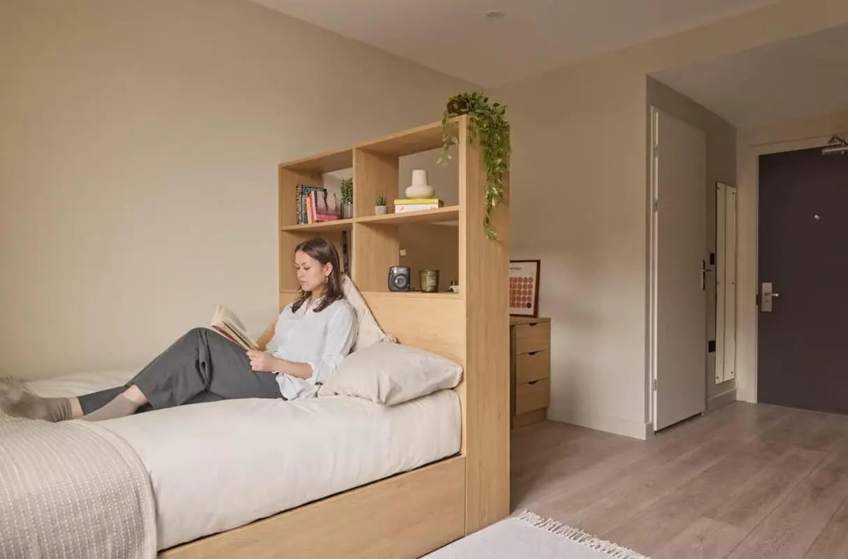 Woman reading on bed with bookshelf bedroom at Ark Canning Town in London Student Accommodation