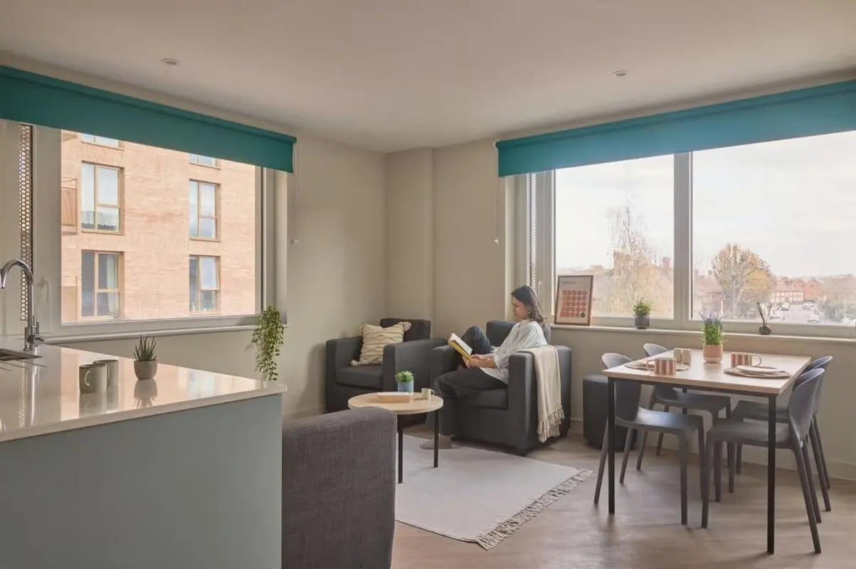Woman reading in armchair dining table and kitchen living room at Ark Canning Town in London Student Accommodation