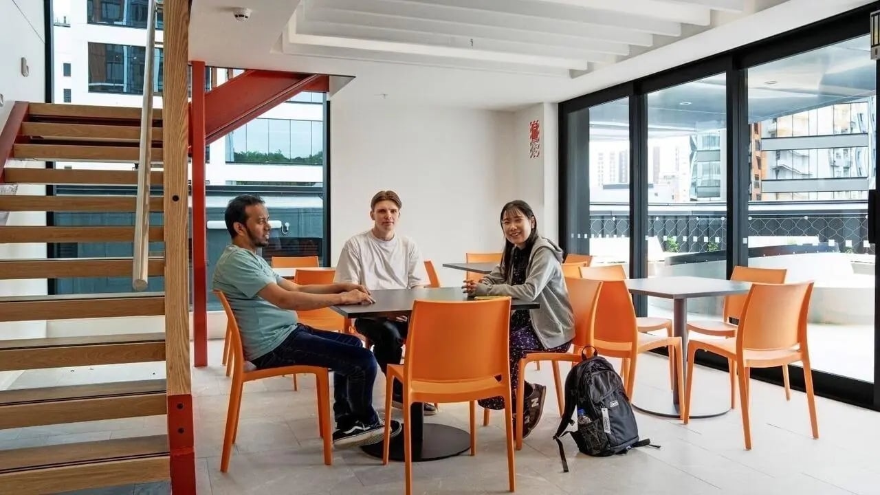 Three students sit at tables near stairs communal area at UniLodge Auckland Central in Auckland Student Accommodation