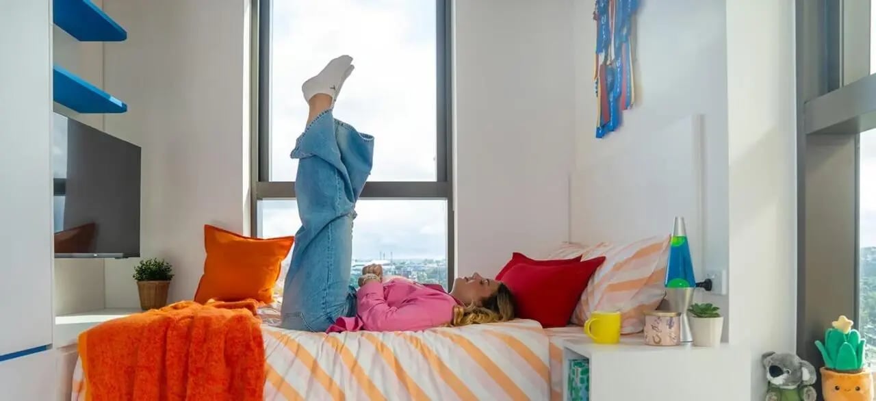 Woman relaxing on bed with orange blanket and pillows bedroom at Scape Kensington in Sydney Student Accommodation