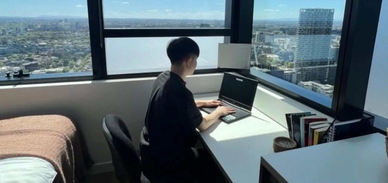 Student working on laptop beside city view bedroom at UniLodge Melbourne Flagstaff in Melbourne Student Accommodation